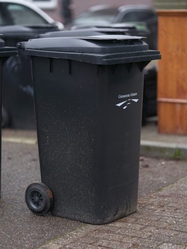 Schwarze Mülltonne mit Deckel auf einem gepflasterten Weg.