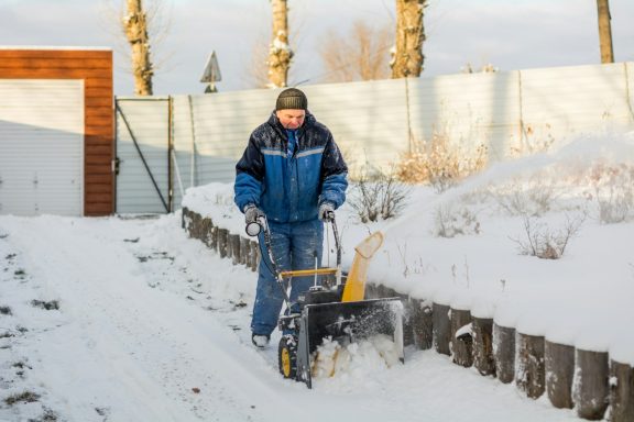 Mann räumt Schnee mit Schneefräse in einem winterlichen Garten.